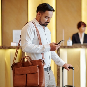Traveler with a leather bag and suitcase using a smartphone in a hotel or airport lobby, searching for the best credit card with airport lounge access.